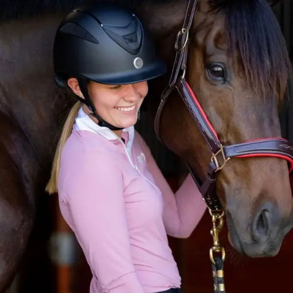 Woman in a pink shirt and helmet standing next to a brown horse.