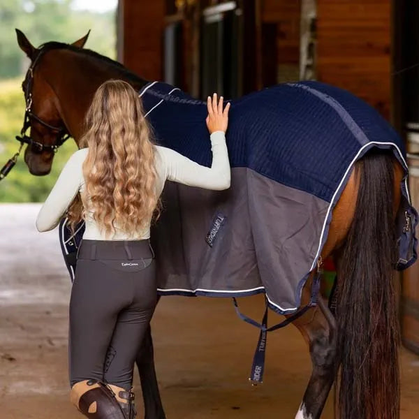 Woman adjusting a blue and gray saddle blanket on a horse in an indoor setting.